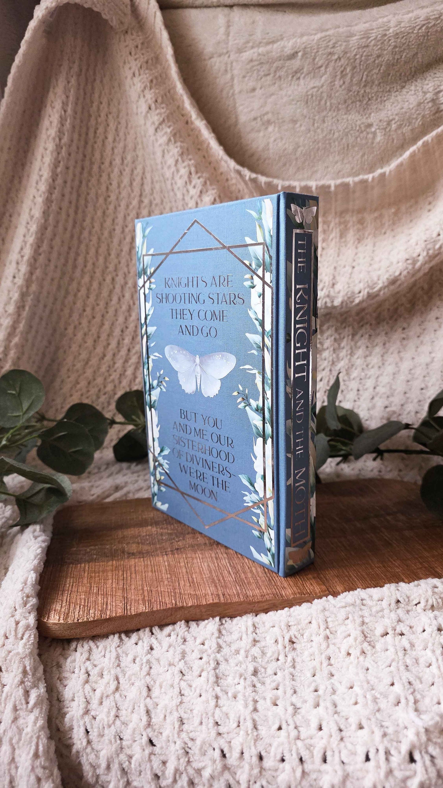 A hardcover book titled 'The Knight and the Moon' with a light blue cover and white butterfly illustration, standing upright on a wooden board surrounded by a beige knit blanket and green leaves.