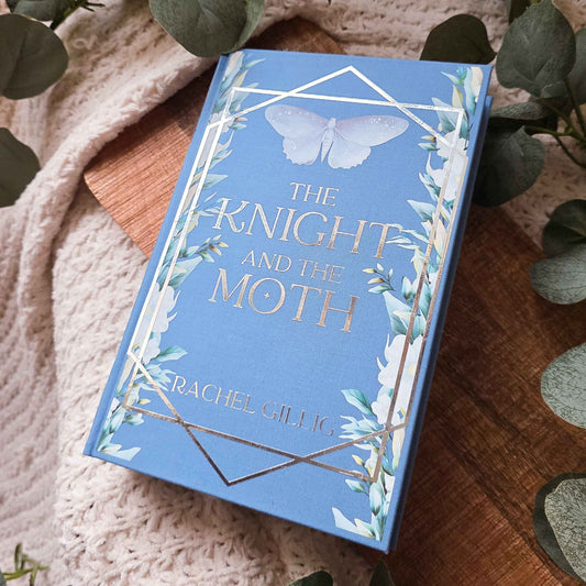 A blue book titled 'The Knight and the Moth' by Rachel Gift 16 lies on a wooden board surrounded by a beige knit blanket and green eucalyptus leaves.