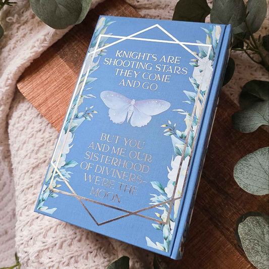 A blue hardcover book titled 'Knights Are Shooting Stars They Come And Go' lies on a wooden board surrounded by green eucalyptus leaves and a beige knit blanket.