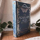 The book 'The Knight and the Moth' by Rachel Cruz stands upright on a wooden tray, surrounded by cozy beige knit blankets and green eucalyptus leaves.