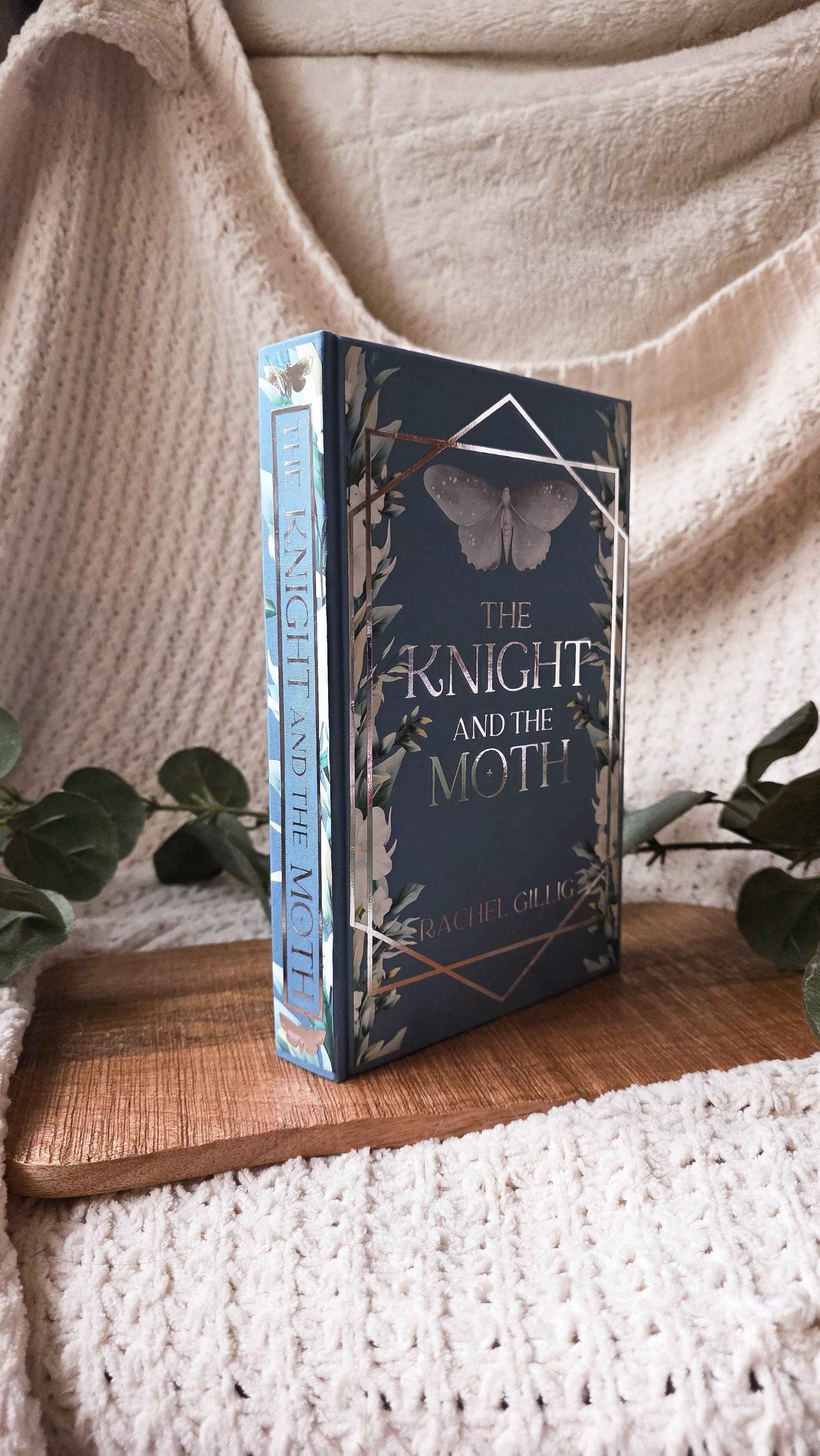 The book 'The Knight and the Moth' by Rachel Cruz stands upright on a wooden tray, surrounded by cozy beige knit blankets and green eucalyptus leaves.