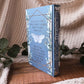 A hardcover book titled 'The Knight and the Moon' with a light blue cover and white butterfly illustration, standing upright on a wooden board surrounded by a beige knit blanket and green leaves.