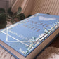 A hardcover book titled 'The Knight and the Moth' by Rachel Gillig, featuring a light blue cover with white butterfly and moth illustrations, placed on a textured cream knit blanket near a window.