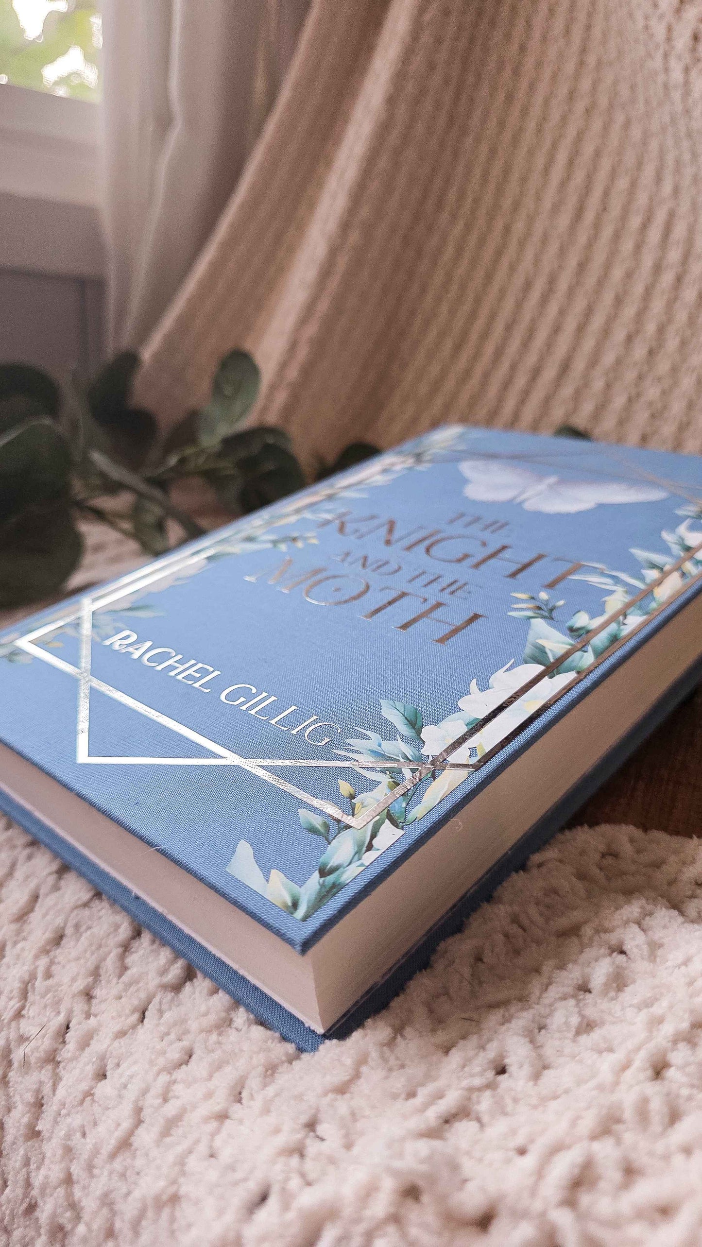 A hardcover book titled 'The Knight and the Moth' by Rachel Gillig, featuring a light blue cover with white butterfly and moth illustrations, placed on a textured cream knit blanket near a window.