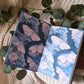 An open notebook or journal lying on a cream knit blanket, decorated with green eucalyptus leaves. The left page features pink butterflies on a dark background, and the right page features pale pink butterflies on a light blue background.