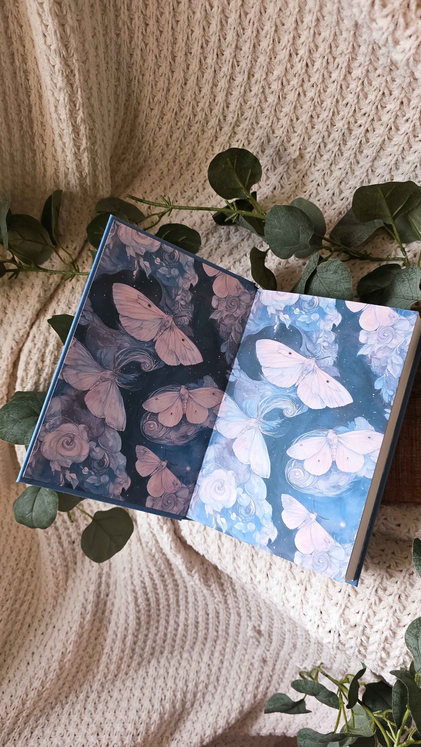 An open notebook or journal lying on a cream knit blanket, decorated with green eucalyptus leaves. The left page features pink butterflies on a dark background, and the right page features pale pink butterflies on a light blue background.