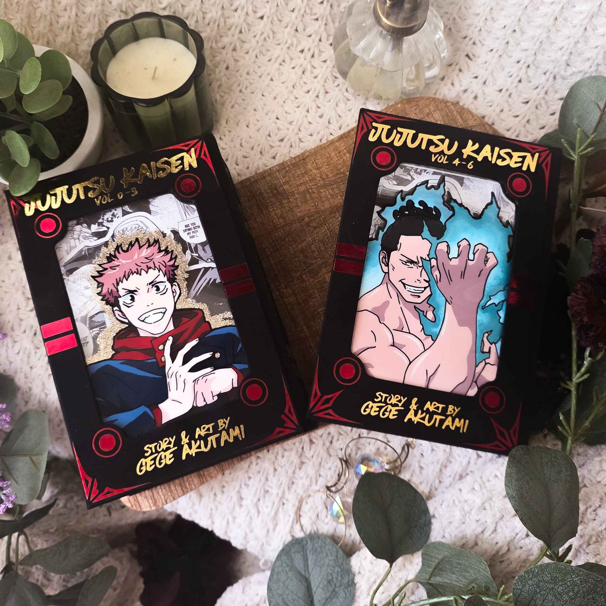 Two illustrated product boxes for 'Jujutsu Kaisen' by Osuke Meltanii are displayed on a wooden board surrounded by decorative plants, a candle, and a soap dispenser against a textured knit background.