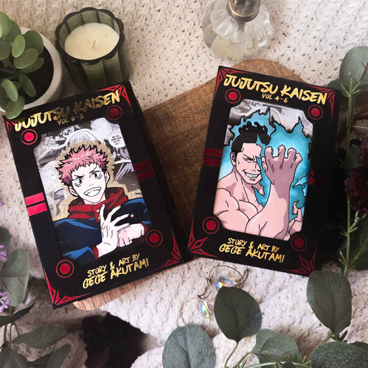 Two illustrated product boxes for 'Jujutsu Kaisen' by Osuke Meltanii are displayed on a wooden board surrounded by decorative plants, a candle, and a soap dispenser against a textured knit background.
