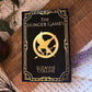 A styled flat-lay of the book 'The Hunger Games' by Suzanne Collins, featuring a black cover with gold lettering and the iconic mockingjay logo, placed on a wooden cutting board over a cream knit blanket with green leaves and dried flowers.