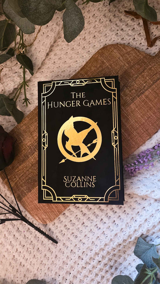 A styled flat-lay of the book 'The Hunger Games' by Suzanne Collins, featuring a black cover with gold lettering and the iconic mockingjay logo, placed on a wooden cutting board over a cream knit blanket with green leaves and dried flowers.